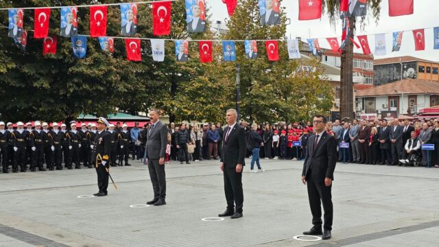 Tören alanında üç sivil yetkili ve bir subay duruyor; arkada bayraklar, denizci askerler ve izleyiciler görünüyor.