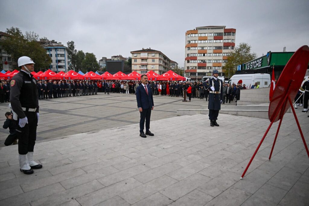Meydanda takım elbiseli bir yetkili çelenk önünde duruyor; yanında nöbetçi askerler, arkada kırmızı çadırlar ve kalabalık var.