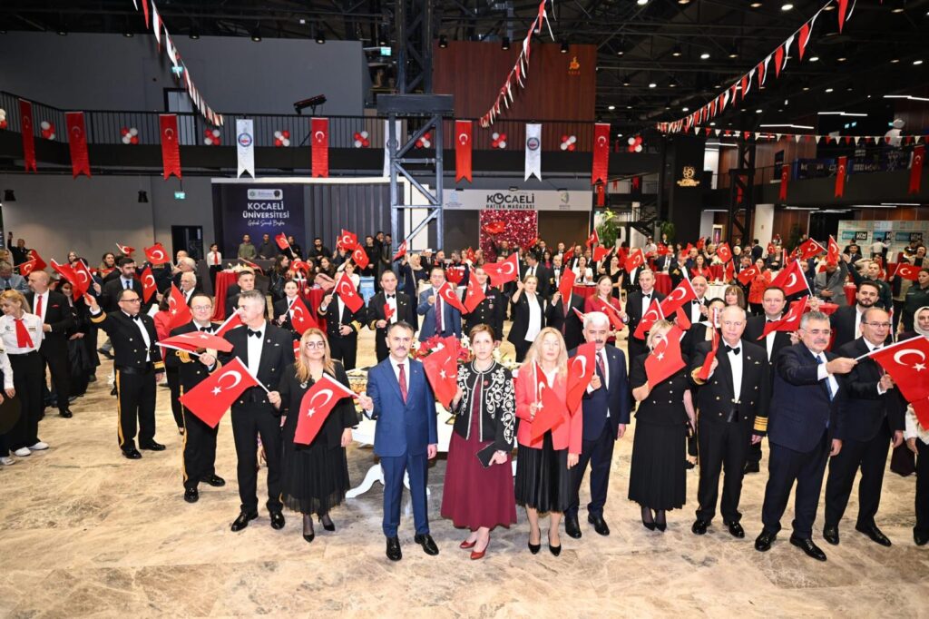 Kongre salonunda, Türk bayrakları sallayan resmi giyimli kadın ve erkekler kameraya poz veriyor; arkada kalabalık ve bayraklar var.