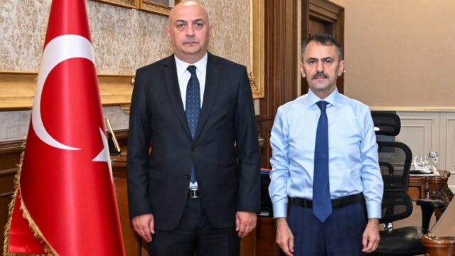 Makam odasında Türk bayrağının yanında koyu takım elbiseli ve açık mavi gömlekli iki erkek yan yana poz veriyor.