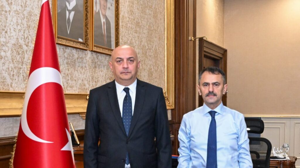 Makam odasında Türk bayrağının yanında koyu takım elbiseli ve açık mavi gömlekli iki erkek yan yana poz veriyor.