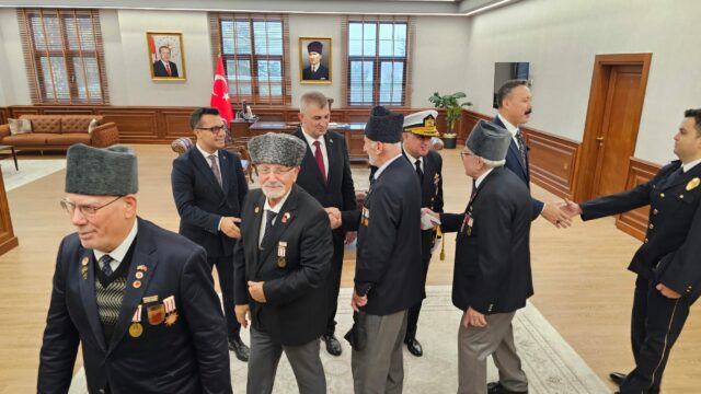 Makam odasında kalpaklı, madalyalı yaşlı erkekler sırayla yetkililerle tokalaşıyor.