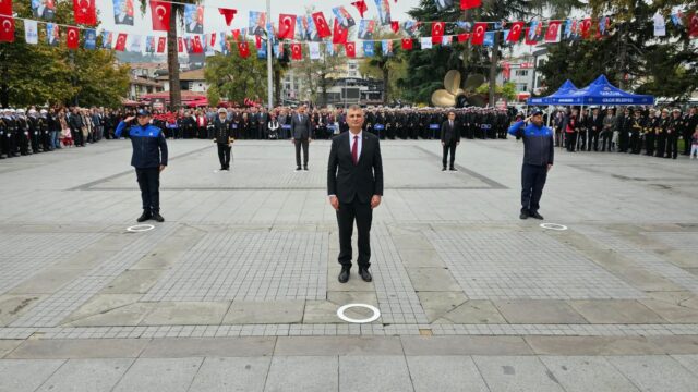 Takım elbiseli bir adam meydanın ortasında dururken arkada yetkililer, askerler ve bayraklarla süslü kalabalık var.