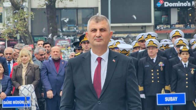 Koyu takım elbiseli, kırmızı kravatlı bir adamın yakın çekimi; arkada üniformalı donanma personeli ve kalabalık var.