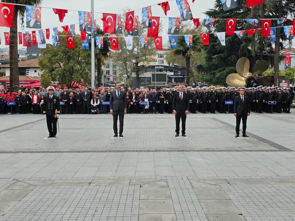 Üç sivil yetkili ve üniformalı bir subay meydanda sıra halinde duruyor, arkada tören kalabalığı ve bayraklar var.