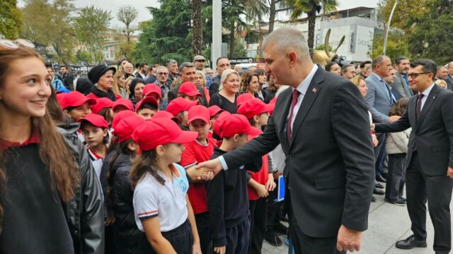Takım elbiseli bir adam kırmızı şapkalı okul çocuklarıyla tokalaşıyor; solda gülümseyen bir genç kız görünüyor.