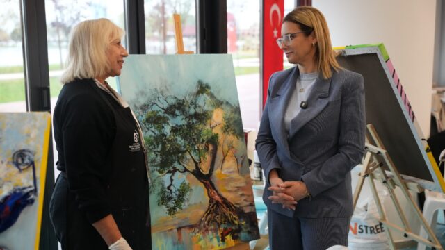 Mavi blazerlı sarışın bir kadın, siyah önlüklü bir sanatçıyla ağaç resmi önünde sohbet ediyor.
