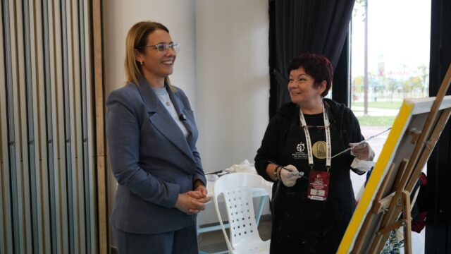 Gri blazerlı gülümseyen bir kadın, şövale başındaki önlüklü sanatçıyla konuşuyor.