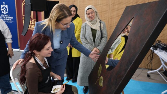 Mavi takımlı bir kadın, oturan genç sanatçının çalışmasına bakıyor; arkada başörtülü kadınlar izliyor.