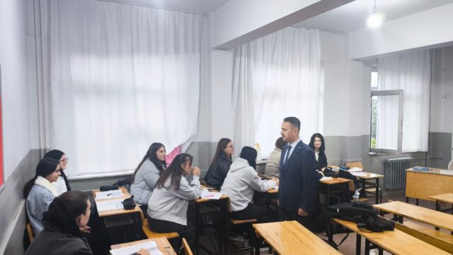 Sınıfta sıralarda oturan öğrencilere takım elbiseli bir erkek bakıyor; pencerelerde beyaz perdeler var.