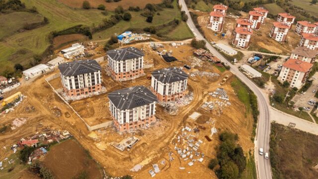 İnşaat halindeki lojman binalarının havadan görüntüsü; sağda kırmızı çatılı tamamlanmış konutlar ve yol görünüyor