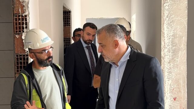 İnşaat içindeki koridorda takım elbiseli bir yetkili, baret takan bir çalışanı dinliyor; arkada başka kişiler var