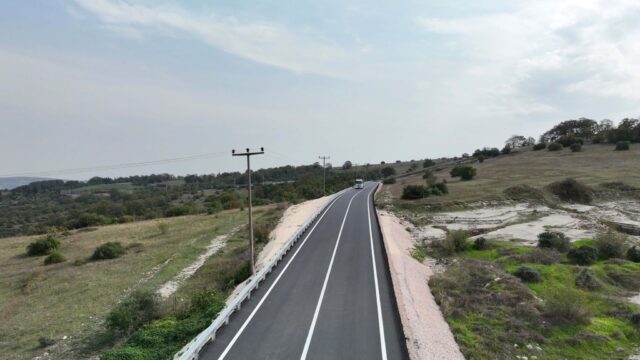 Bir kısmında bariyer bulunan, yeni asfaltlı ve beyaz şeritli yolda uzaktaki bir araç ile elektrik direkleri.