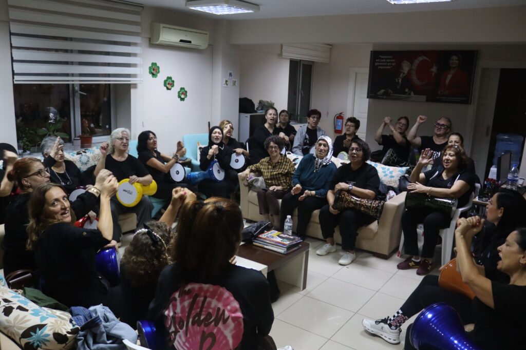 Siyah giysili kadınlar bir odada koltuklarda oturup darbuka ve def çalıyor, bazıları gülümseyip el çırpıyor.