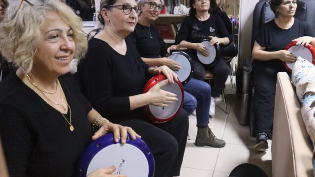 Siyah giyen gözlüklü kadınlar ellerindeki darbukalara vuruyor; öndeki sarışın kadın gülümsüyor.