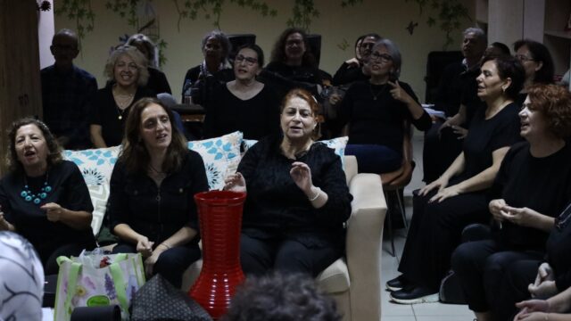 Siyah giyen kadınlar koltuklarda oturup şarkı söylüyor; önde kırmızı bir vazo ve alışveriş poşeti var.