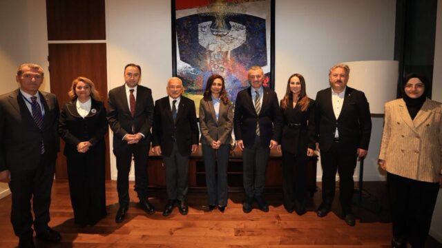 Duvarda büyük bir tablonun önünde, resmi giyimli sekiz kişilik grup kameraya bakarak poz veriyor