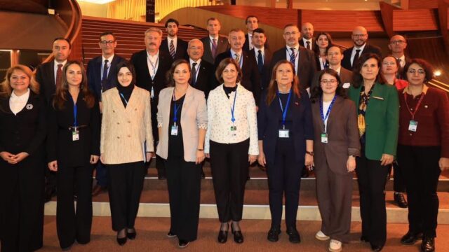 Salonda merdivenlerin önünde toplu fotoğraf için poz veren, yakalarında kimlik kartı olan takım elbiseli kadın ve erkek heyet üyeleri