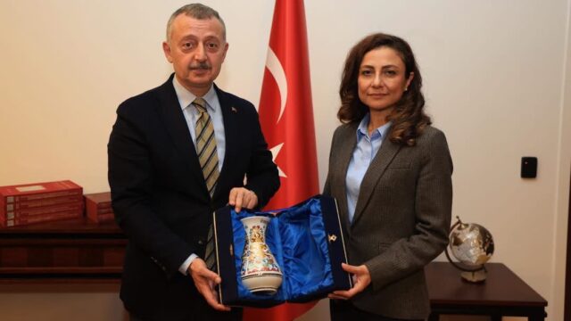 Türk bayrağının önünde, takım elbiseli bir erkek ve gri ceketli bir kadın mavi kutu içindeki vazoyu birlikte tutuyor