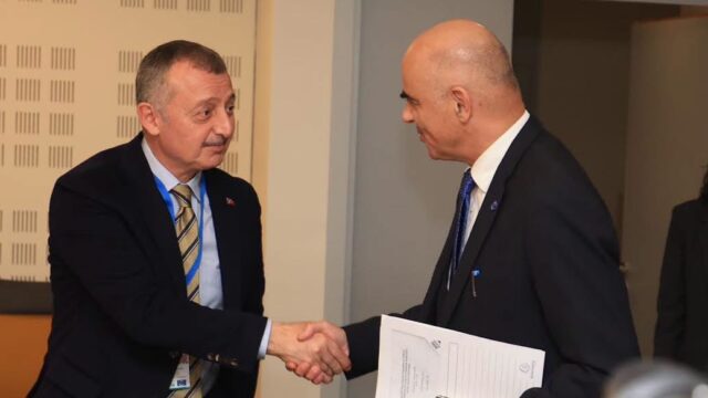 Takım elbiseli iki erkek elde belge tutarken tokalaşıyor, arkada bir kadın görünüyor