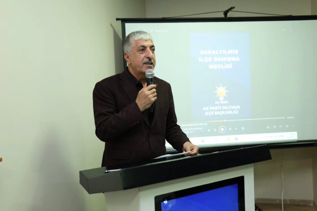 Kürsüde mikrofonla konuşan koyu ceketli, gri saçlı bir adam; arkasında AK Parti Dilovası İlçe Başkanlığı yazılı ekran var.