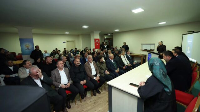 Toplantı salonunda sıralara oturmuş kalabalık dinleyici grubu; sağ arkada ayakta konuşan bir kişi ve ekran görünüyor.