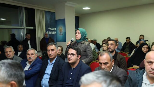 Dinleyiciler arasında ayakta mikrofonla konuşan yeşil başörtülü bir kadın; çevresinde oturan erkekler ve kadınlar var.