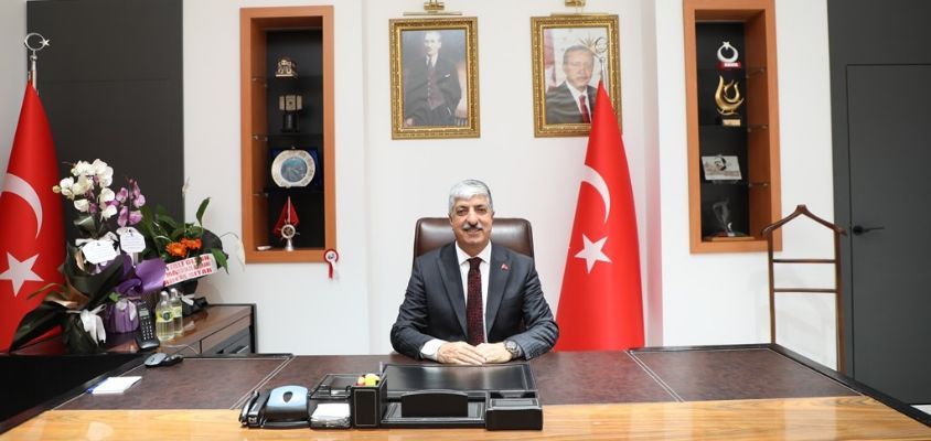 Makam odasında masasının arkasında oturan gri saçlı, koyu takım elbiseli bir adam; yanlarda Türk bayrakları ve çiçekler var.