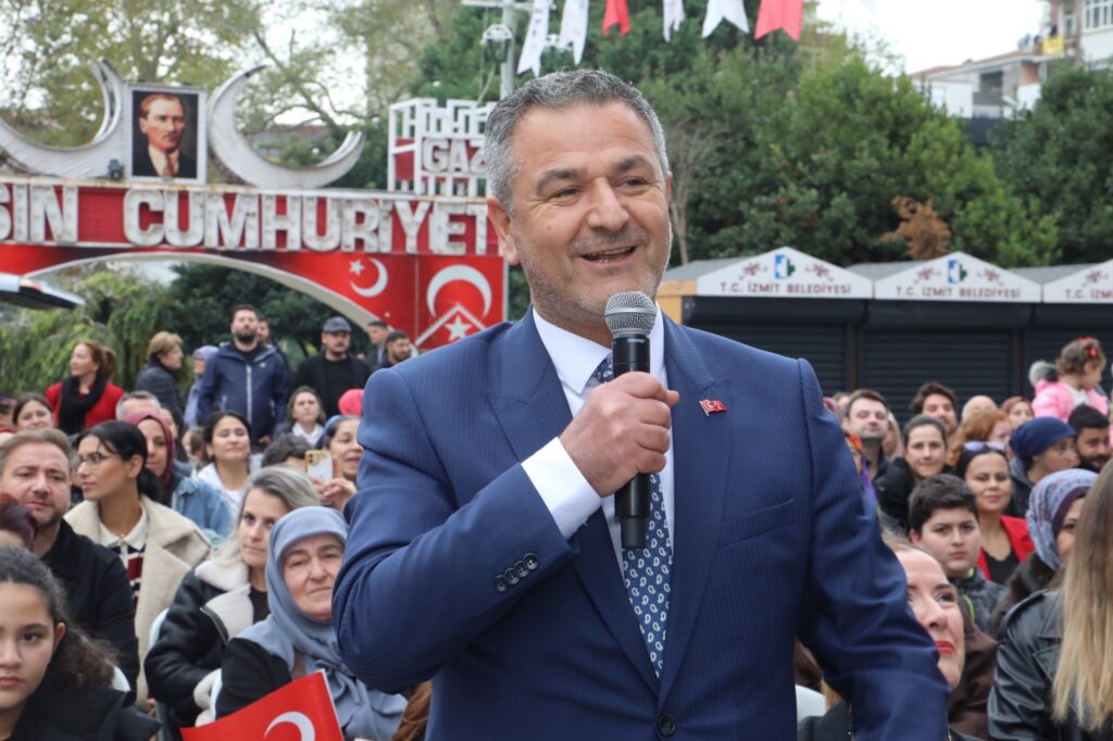 Lacivert takım elbiseli bir adam mikrofonla konuşuyor; arkasında Cumhuriyet yazılı süs ve kalabalık izleyici var.