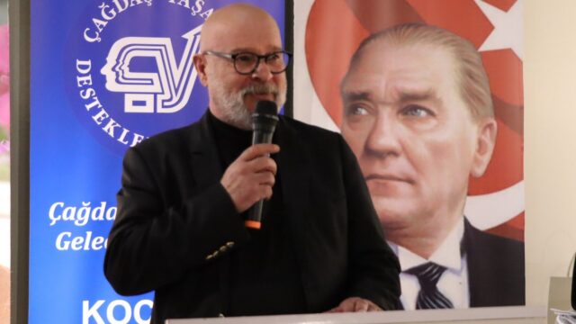 Siyah giyimli, gözlüklü sakallı bir adam mikrofonla konuşuyor; arkasında ÇYDD pankartı ve Türk bayraklı Atatürk portresi var.
