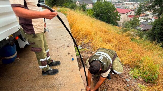 Yamaçta bir işçi siyah boruyu tutarken diğeri çamurlu çukurda su hattını onarıyor.