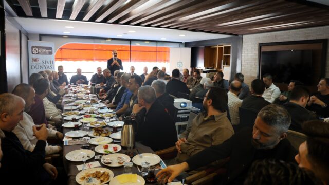 Yemekli toplantıda masalara oturan esnaf, ayakta mikrofonla konuşan bir kişiyi dinliyor