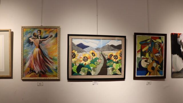 Galeri duvarında yan yana asılı renkli tablolar: dans eden çift, ayçiçekleri ve soyut bir kompozisyon.