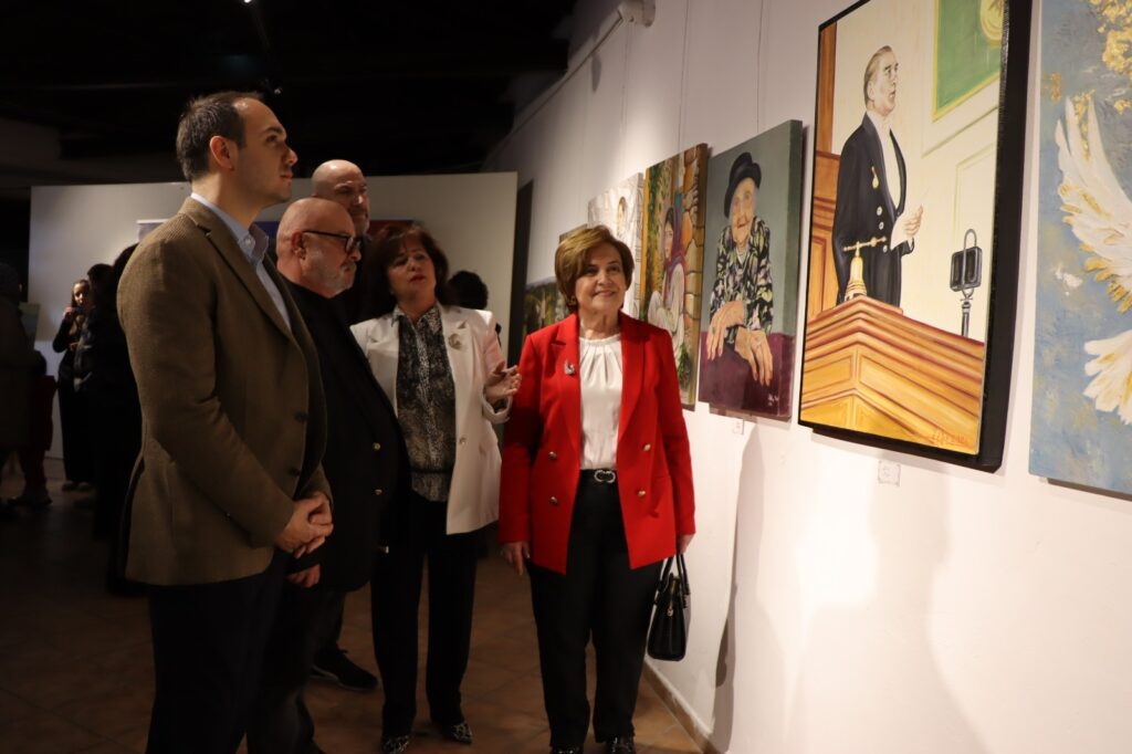 Kırmızı ceketli kadın ve diğer ziyaretçiler galeride duvardaki tablolara bakıyor.