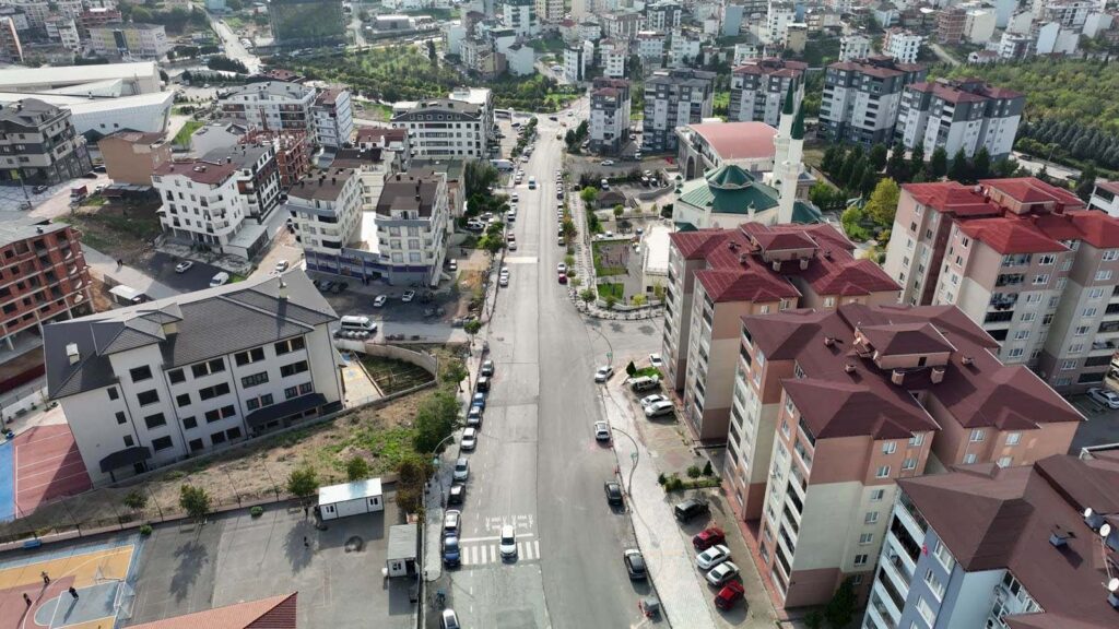 Havadan çekilmiş, iki yanında apartmanlar ve bir caminin bulunduğu uzun, düz cadde ile park etmiş araçlar.