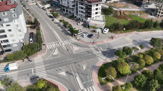 Havadan görünen, yaya geçitleri olan geniş bir kavşak, çevresinde apartmanlar, ağaçlar ve park halindeki araçlar.