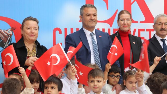 Takım elbiseli bir adam ve yanındaki yetişkinler, önlerinde bayrak sallayan çocuklarla kameraya bakıyor.