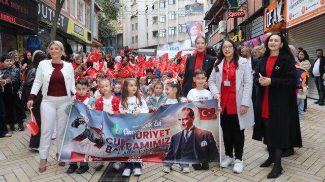 Çocuklar ve yetişkinler Cumhuriyet Bayramı pankartını taşıyarak caddede yürüyor, arkada bayraklı kalabalık var.