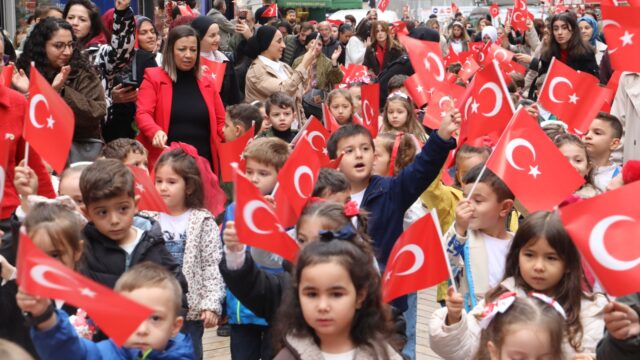 Türk bayrağı sallayan çocuklar caddede yürüyor, kenarda alkışlayan yetişkinler izliyor.