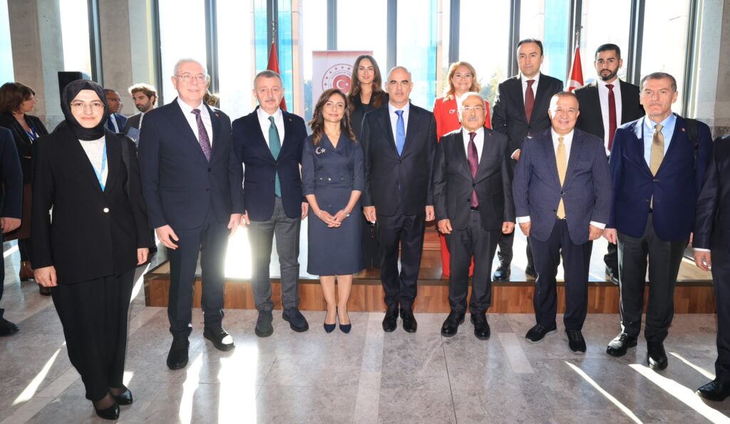 Resepsiyonda takım elbiseli erkekler ve kadınlar, arkasında Türk bayrakları olan salonda toplu fotoğraf için poz veriyor.