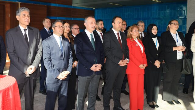 Takım elbiseli erkekler ve kırmızı takımlı bir kadın, resepsiyonda ayakta bir konuşmayı dinliyor.