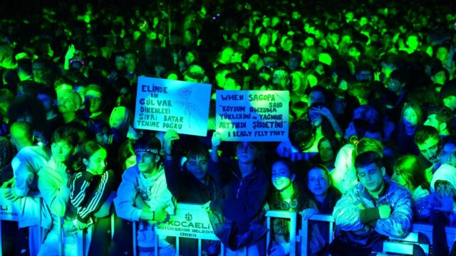 Yeşil ışık altında kalabalık konser izleyicileri, bariyer önünde yazılı iki pankart tutuyor.