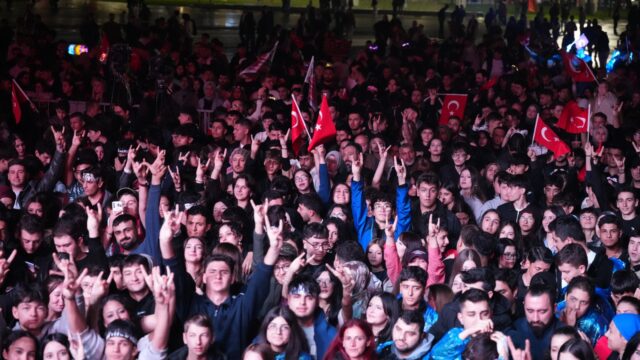 Gece meydanda kalabalık, birçok kişi elleriyle işaret yapıyor, aralarında Türk bayrakları var.