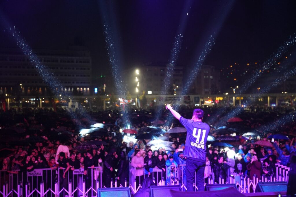41 numaralı formalı sanatçı sahnede kolunu kaldırıyor, önünde şemsiyeli kalabalık, ışık huzmeleri var.