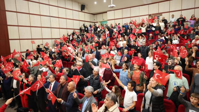 Dolu salonda seyirciler ellerinde Türk bayraklarını sallıyor; aralarında çocuklar da var.
