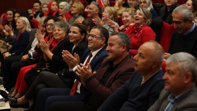 Salondaki seyirciler gülümseyerek alkışlıyor; ön sırada takım elbiseli gözlüklü bir adam görülüyor.