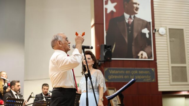 Beyaz gömlekli yaşlı bir adam mikrofonla konuşuyor; yanında mavi elbiseli kadın var, arkada Atatürk portresi asılı.