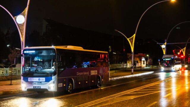Gece ıslak caddede ilerleyen mor beyaz şehirlerarası otobüs; arkada bir başka otobüs ve sokak lambaları var.