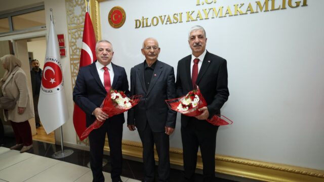 Üç adam, ikisi çiçek buketi tutarak, Dilovası Kaymakamlığı yazılı duvarın önünde kameraya poz veriyor.