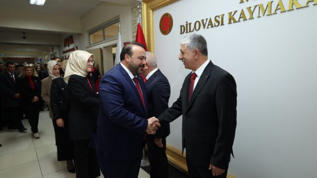 Lacivert takım elbiseli bir adam, kaymakamlık binasında gri saçlı bir adamla tokalaşıyor; arkada sıra bekleyenler var.